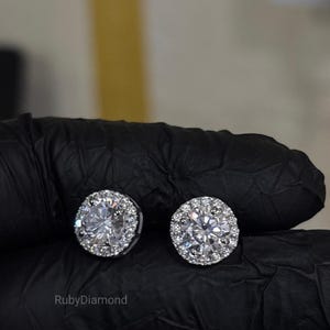 May include: A pair of round stud earrings featuring a large central clear stone surrounded by a halo of smaller clear stones. The earrings are set in a metallic frame. The text RubyDiamond is visible in the lower left corner of the image.