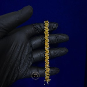 May include: A tennis bracelet featuring a single row of pear-cut yellow gemstones set in a metallic frame. The bracelet is held vertically against a dark blue background, showcasing the uniform shape and vibrant color of each individual stone.