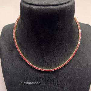 Puede incluir: Un collar de tenis en tono dorado que presenta una fila continua de pequeñas piedras redondas de color rojo vibrante. El collar se muestra sobre un busto negro y el texto RubyDiamond está impreso en blanco en la parte inferior izquierda.