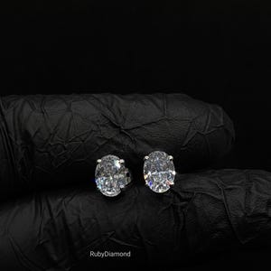 Peut inclure: Une paire de boucles d'oreilles à clous ovales transparentes avec des montures en métal argenté. Les boucles d'oreilles présentent une surface facettée qui reflète la lumière, créant un aspect brillant. Le texte RubyDiamond est inscrit en blanc.