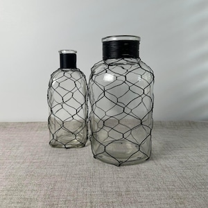May include: Two clear glass bottles of different sizes, each wrapped in a black wire mesh netting. The necks of both bottles are tightly wound with black wire, creating a uniform look for these decorative glass containers.