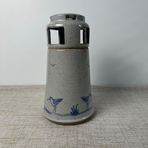 May include: A speckled light gray ceramic lighthouse-shaped candle holder with a dark blue hand-painted design featuring two shorebirds and a small plant. The structure has a cylindrical top section with four rectangular cutouts for light.