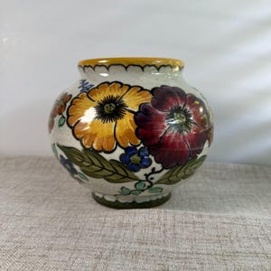 May include: A round ceramic vase with a cream-coloured base featuring a hand-painted floral pattern. The design includes large yellow and deep red flowers, smaller blue blossoms, and green leaves. The rim is accented with a yellow band.