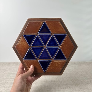 May include: A hexagonal ceramic tile featuring a geometric pattern of dark blue glazed triangles arranged in a central star shape, surrounded by larger, unglazed, earthy brown triangular and trapezoidal sections with a textured finish.