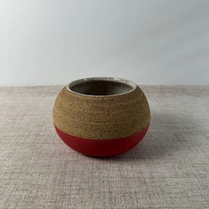 May include: A spherical ceramic bowl featuring a two-tone design with a speckled tan upper section and a solid red lower section. The exterior surface of the tan portion displays fine horizontal ridges, while the interior is light and speckled.