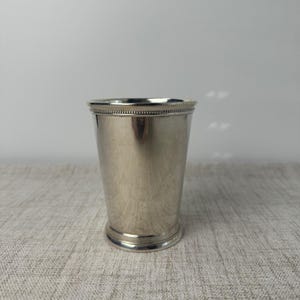 May include: A silver-toned metal tumbler featuring a smooth, tapered body and a decorative beaded rim detail at the top edge and the base. The cup has a polished, reflective surface and a simple, functional design with a wide opening.