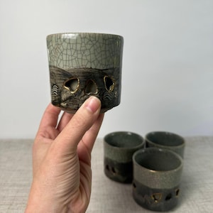 May include: A set of ceramic cups featuring a two-tone design with a light green crackle glaze on the upper portion and a dark, textured base. Each cup includes small, heart-shaped cutouts along the middle, accented with gold-colored trim.