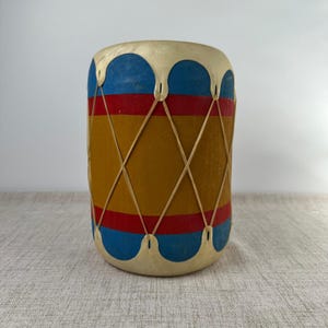 May include: A cylindrical drum featuring a central mustard yellow band bordered by red stripes and blue scalloped edges at the top and bottom. The drum is wrapped in a crisscross pattern of thin, light-colored cord lacing across the body.