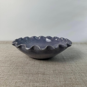 May include: A shallow, circular ceramic bowl with a glossy, muted purple glaze. The rim of the bowl features a distinct, wavy, ruffled edge that creates a scalloped appearance around the entire circumference of the dish.