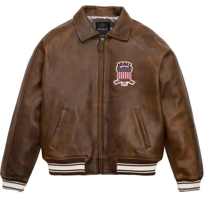 K&ouml;nnte beinhalten: Braune Lederjacke mit Rei&szlig;verschluss, Kragen und Brustaufn&auml;her. Die Jacke hat wei&szlig;-schwarz gestreifte B&uuml;ndchen und Bund. Der Aufn&auml;her zeigt ein Schilddesign mit dem Text "AVIREX".