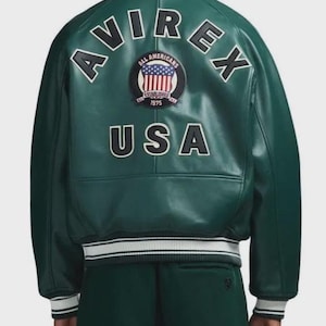 May include: A dark green leather jacket with white and black accents. The back of the jacket features the word "AVIREX" in white block letters, along with a patch that says "ALL AMERICANS" and "USA". The jacket has a ribbed collar and cuffs.
