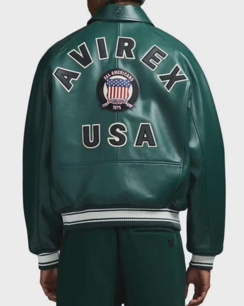 May include: A dark green leather jacket with white and black accents. The back of the jacket features the word "AVIREX" in white block letters, along with a patch that says "ALL AMERICANS" and "USA". The jacket has a ribbed collar and cuffs.