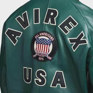 May include: Green leather jacket with white and black lettering spelling out "AVIREX" and "USA". A circular patch features a shield with the American flag and the text "ALL AMERICANS ESTABLISHED 1975 N.Y."