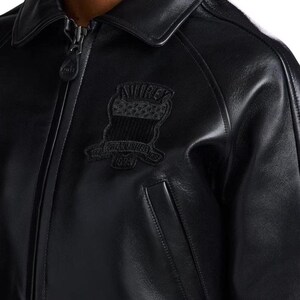 Op de afbeelding: Zwarte leren jas met een kraag en een ritssluiting. De jas heeft een patch op de borst met de tekst "AVIREX" en "ESTABLISHED 1975". De jas heeft een klassiek ontwerp met een zak aan de voorkant.