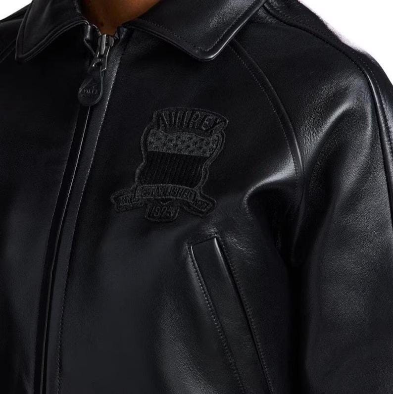 Op de afbeelding: Zwarte leren jas met een kraag en een ritssluiting. De jas heeft een patch op de borst met de tekst "AVIREX" en "ESTABLISHED 1975". De jas heeft een klassiek ontwerp met een zak aan de voorkant.