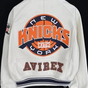 May include: A white leather varsity jacket featuring a large back graphic with an orange basketball and the text NEW YORK KNICKS KITH. The word AVIREX is embroidered below in dark lettering. The jacket has a ribbed collar and striped cuffs.