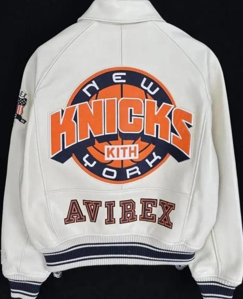 May include: A white leather varsity jacket featuring a large back graphic with an orange basketball and the text NEW YORK KNICKS KITH. The word AVIREX is embroidered below in dark lettering. The jacket has a ribbed collar and striped cuffs.