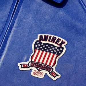 May include: A bright blue leather jacket featuring an embroidered patch. The patch displays a shield with red and white stripes and a blue field with white stars. Text on the patch reads AVIREX, ESTABLISHED, 1975, and NY.