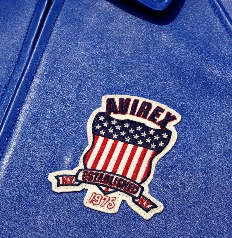 May include: A bright blue leather jacket featuring an embroidered patch. The patch displays a shield with red and white stripes and a blue field with white stars. Text on the patch reads AVIREX, ESTABLISHED, 1975, and NY.