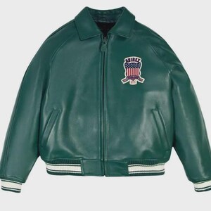 May include: A green leather jacket with a zip-up front, collar, and ribbed cuffs and hem. The jacket features a patch on the chest with a shield design and the text "AVIREX ESTABLISHED 1975". The jacket has two front pockets.