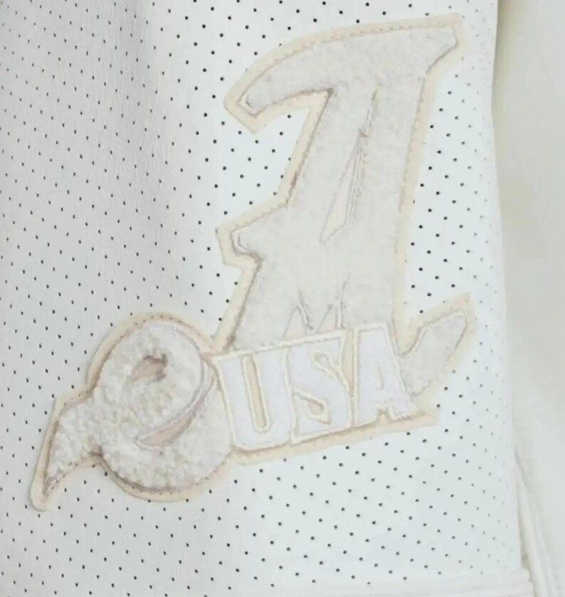 May include: A white perforated garment featuring a large, textured, off-white chenille patch. The patch displays a stylized letter A, a circular emblem, and the text USA in block letters, all stitched onto the surface with a light border.
