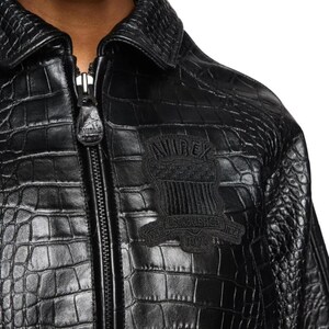May include: A black jacket with a textured crocodile skin pattern, featuring a front zipper closure with a branded pull tab and a black embroidered patch on the chest that reads "AVIREX" and "ESTABLISHED 1975" with a shield graphic.