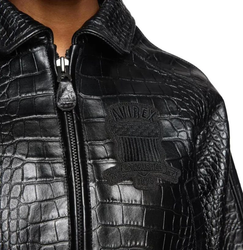May include: A black jacket with a textured crocodile skin pattern, featuring a front zipper closure with a branded pull tab and a black embroidered patch on the chest that reads "AVIREX" and "ESTABLISHED 1975" with a shield graphic.