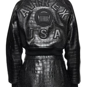 May include: A black jacket and matching skirt set featuring a crocodile-embossed texture. The back of the jacket displays the text AVIREX USA in large letters with a central circular emblem featuring a shield and stars design in black.