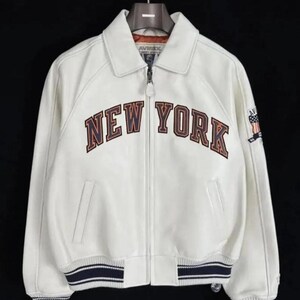 May include: A white leather jacket featuring a full-length front zipper, a classic collar, and ribbed cuffs and hem with navy blue stripes. The chest displays the text NEW YORK in dark blue and orange lettering, with an American flag patch on the sleeve.