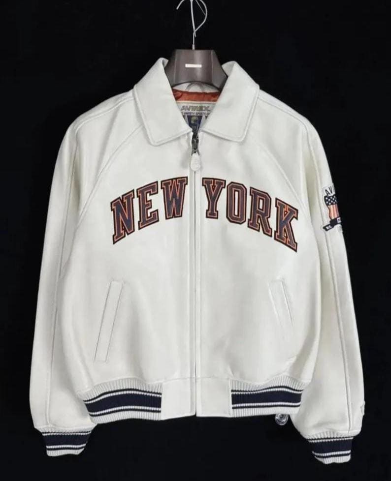 May include: A white leather jacket featuring a full-length front zipper, a classic collar, and ribbed cuffs and hem with navy blue stripes. The chest displays the text NEW YORK in dark blue and orange lettering, with an American flag patch on the sleeve.
