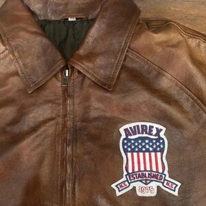 K&ouml;nnte beinhalten: Braune Lederjacke mit Kragen und Rei&szlig;verschluss. Sie hat einen Aufn&auml;her mit dem Text "AVIREX ESTABLISHED 1975 N.Y." und einem Design der amerikanischen Flagge.