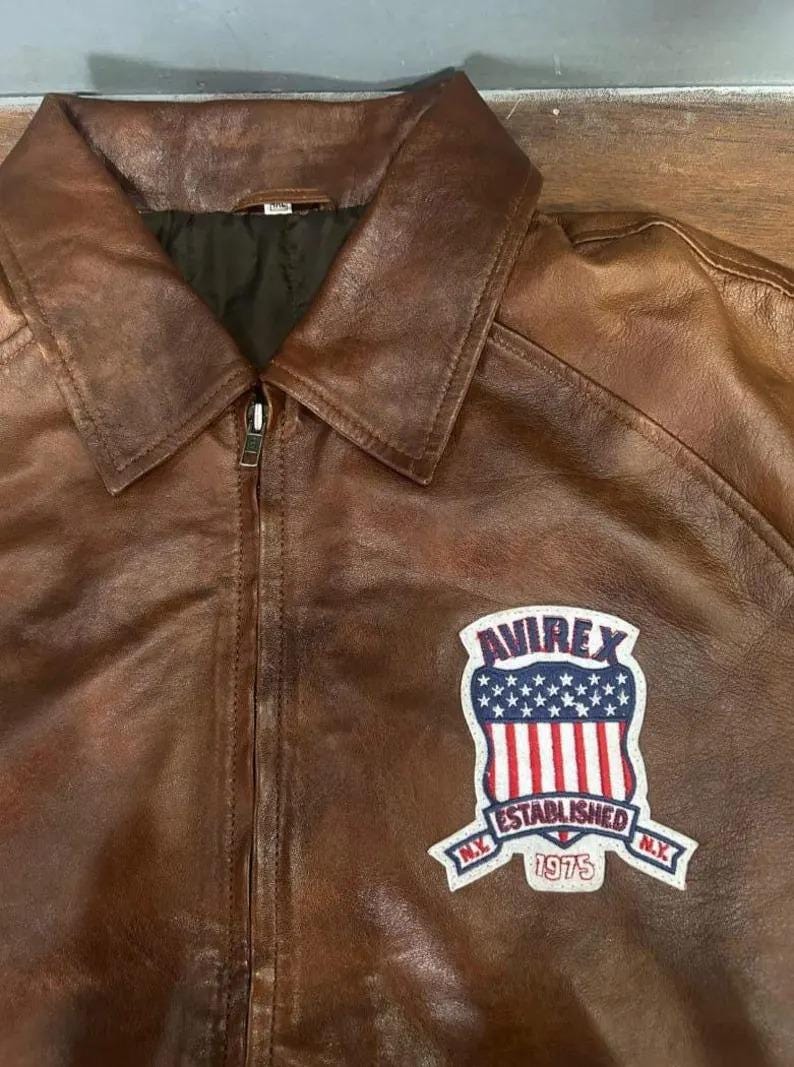 K&ouml;nnte beinhalten: Braune Lederjacke mit Kragen und Rei&szlig;verschluss. Sie hat einen Aufn&auml;her mit dem Text "AVIREX ESTABLISHED 1975 N.Y." und einem Design der amerikanischen Flagge.