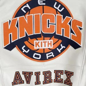 May include: A white jacket featuring a large circular patch on the back with an orange basketball graphic, the text NEW YORK KNICKS, and the word KITH in a white box. Below the patch, the word AVIREX is embroidered in orange and navy blue letters.