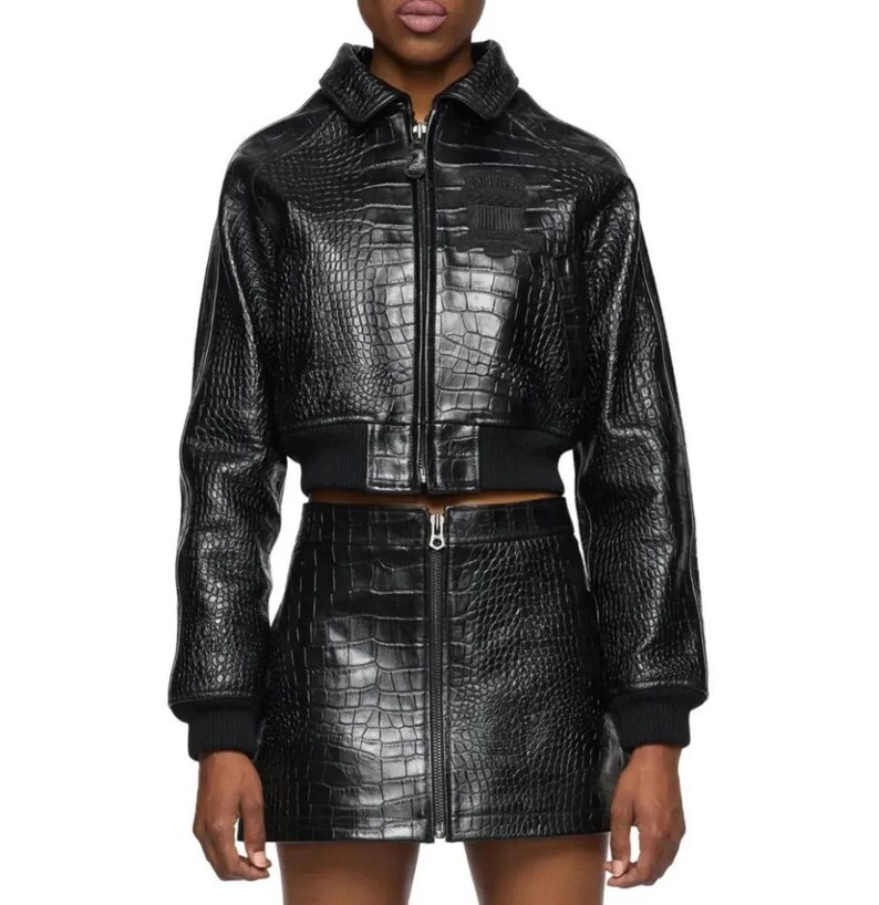 May include: A black cropped jacket and matching mini skirt set featuring a glossy crocodile-embossed texture. The jacket has a collared neckline, a full-length front zipper, and ribbed cuffs. The skirt features a central vertical zipper closure.