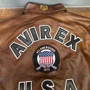 K&ouml;nnte beinhalten: Braune Lederjacke mit dem Wort "AVIREX" in wei&szlig;en Buchstaben mit schwarzer Umrandung. Darunter befindet sich ein Aufn&auml;her mit den Worten "ALL AMERICANS ESTABLISHED 1975" und einem US-Flaggen-Design. Das Wort "USA" steht darunter.