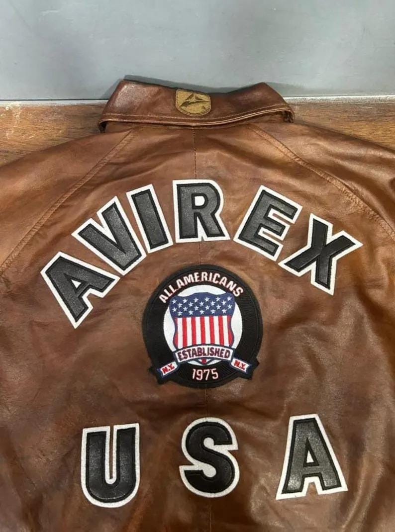 K&ouml;nnte beinhalten: Braune Lederjacke mit dem Wort "AVIREX" in wei&szlig;en Buchstaben mit schwarzer Umrandung. Darunter befindet sich ein Aufn&auml;her mit den Worten "ALL AMERICANS ESTABLISHED 1975" und einem US-Flaggen-Design. Das Wort "USA" steht darunter.