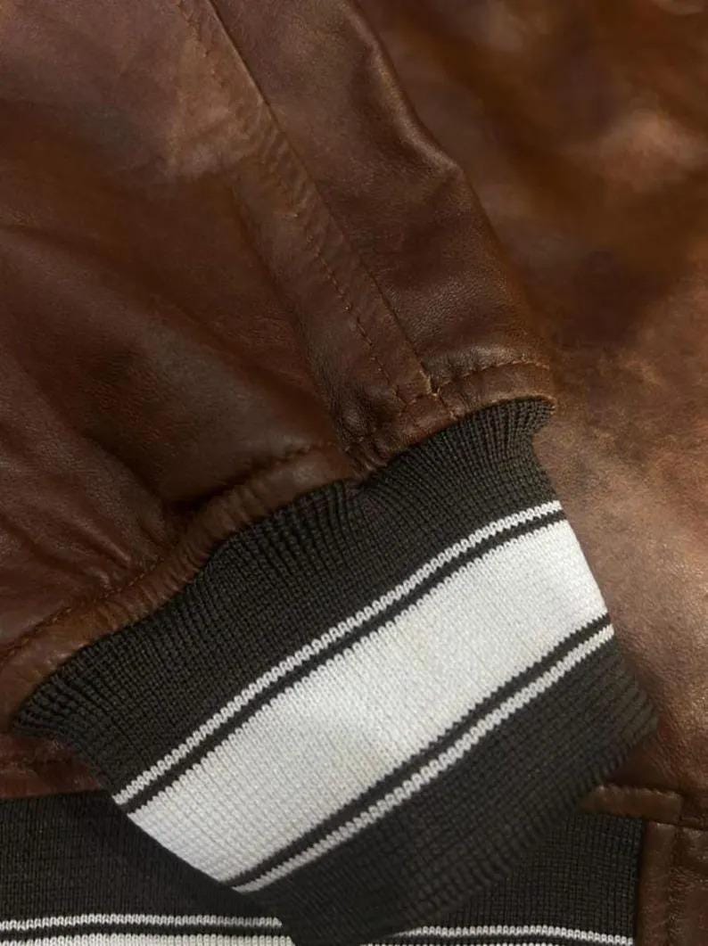 K&ouml;nnte beinhalten: Nahaufnahme eines braunen Lederjacken&auml;rmels. Das B&uuml;ndchen ist schwarz mit zwei wei&szlig;en Streifen. Das Leder hat eine glatte Textur und eine satte, warme Farbe.
