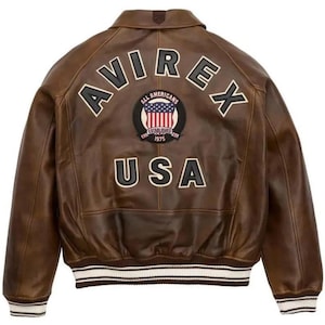 K&ouml;nnte beinhalten: Braune Lederjacke mit dem Wort "AVIREX" in wei&szlig;en Blockbuchstaben &uuml;ber den Schultern. Darunter befindet sich ein Aufn&auml;her mit den Worten "ALL AMERICANS ESTABLISHED 1975 USA" und einer Grafik der amerikanischen Flagge.