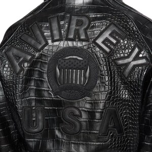 May include: A black jacket with a textured crocodile skin pattern. The back features the word AVIREX in large letters, the word USA below it, and a central circular emblem with a shield and the text ESTABLISHED 1975 and NYC AMERICANS.