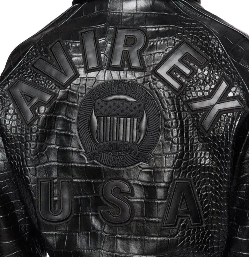 May include: A black jacket with a textured crocodile skin pattern. The back features the word AVIREX in large letters, the word USA below it, and a central circular emblem with a shield and the text ESTABLISHED 1975 and NYC AMERICANS.