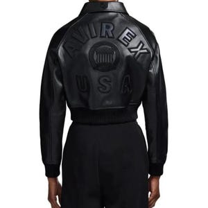 Op de afbeelding: Zwarte leren jas met een bomber-stijl ontwerp. De achterkant heeft een reli&euml;f tekst die "AVIREX USA" leest rond een cirkelvormig embleem. De jas heeft een geribbelde zoom en manchetten en een klassieke kraag.