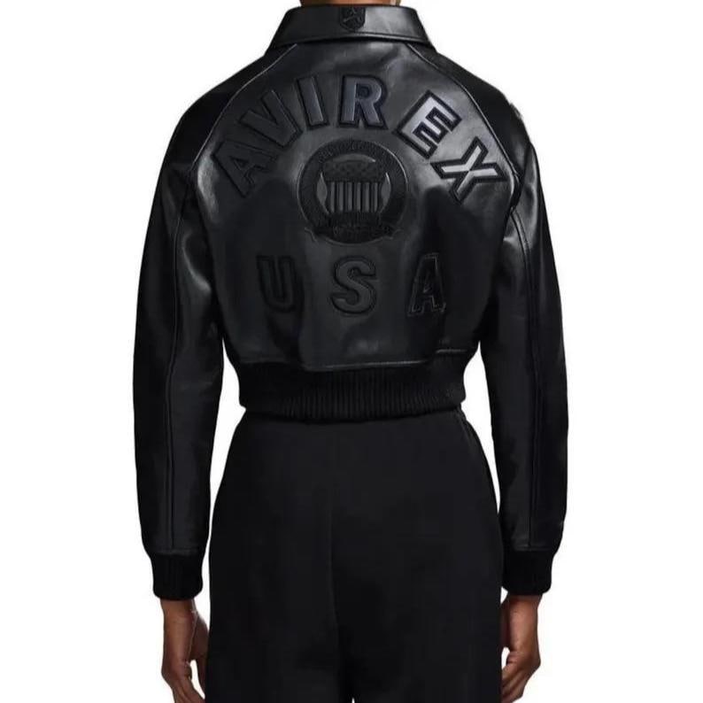 Op de afbeelding: Zwarte leren jas met een bomber-stijl ontwerp. De achterkant heeft een reli&euml;f tekst die "AVIREX USA" leest rond een cirkelvormig embleem. De jas heeft een geribbelde zoom en manchetten en een klassieke kraag.