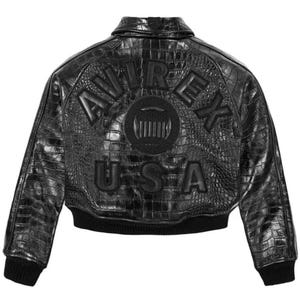 May include: A black jacket with a textured crocodile-embossed finish. The back features the embossed text AVIREX in an arc above a circular emblem and the text USA below. The jacket includes a collar, long sleeves, and a ribbed black hem and cuffs.