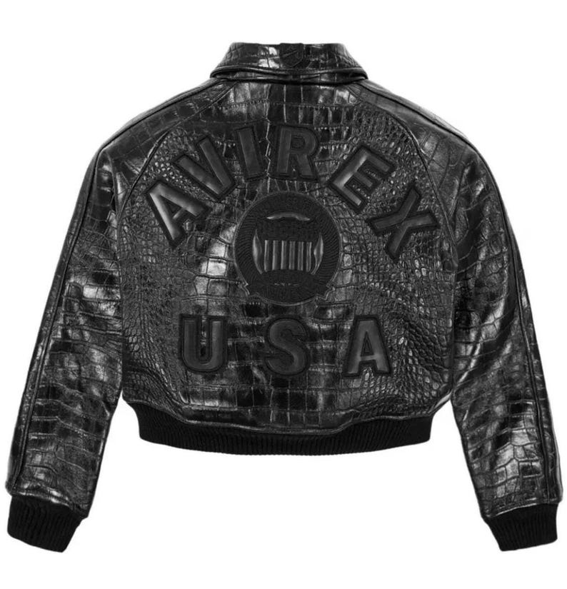 May include: A black jacket with a textured crocodile-embossed finish. The back features the embossed text AVIREX in an arc above a circular emblem and the text USA below. The jacket includes a collar, long sleeves, and a ribbed black hem and cuffs.