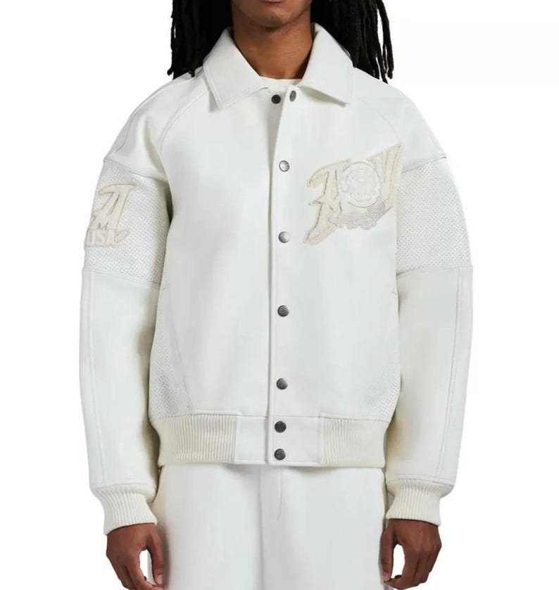 May include: A cream-colored varsity jacket featuring a pointed collar, snap-button front closure, and ribbed cuffs and hem. The jacket displays textured off-white patches on the chest and sleeves, with mesh-like fabric panels on the upper arms.
