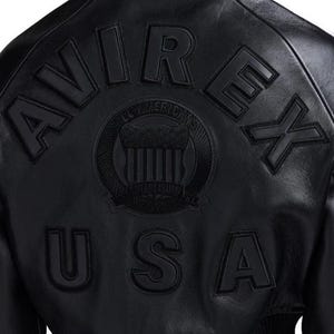 Op de afbeelding: Zwarte leren jas met het woord "AVIREX" in verhoogde letters op de rug. Daaronder staat "USA". Een cirkelvormig embleem is gecentreerd tussen de woorden, met de woorden "ALL AMERICANS" en "ESTABLISHED 1975" rond het embleem.
