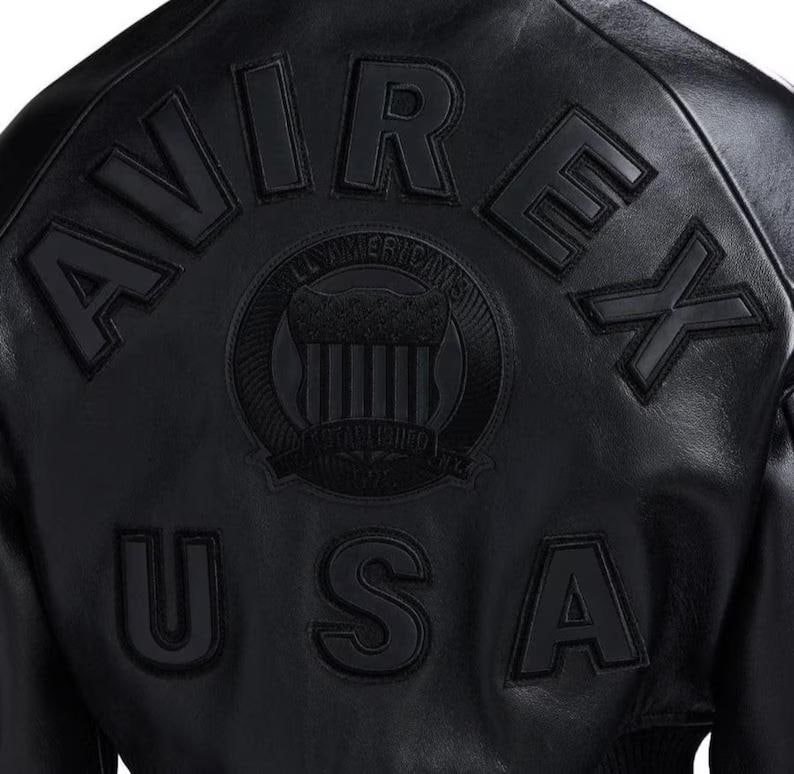 Op de afbeelding: Zwarte leren jas met het woord "AVIREX" in verhoogde letters op de rug. Daaronder staat "USA". Een cirkelvormig embleem is gecentreerd tussen de woorden, met de woorden "ALL AMERICANS" en "ESTABLISHED 1975" rond het embleem.