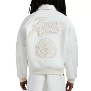 May include: A white varsity jacket featuring a large embroidered Avirex New York logo on the upper back and a circular Avirex Gold Club patch below it. The jacket has a ribbed collar, cuffs, and hem, with textured panels on the sleeves.