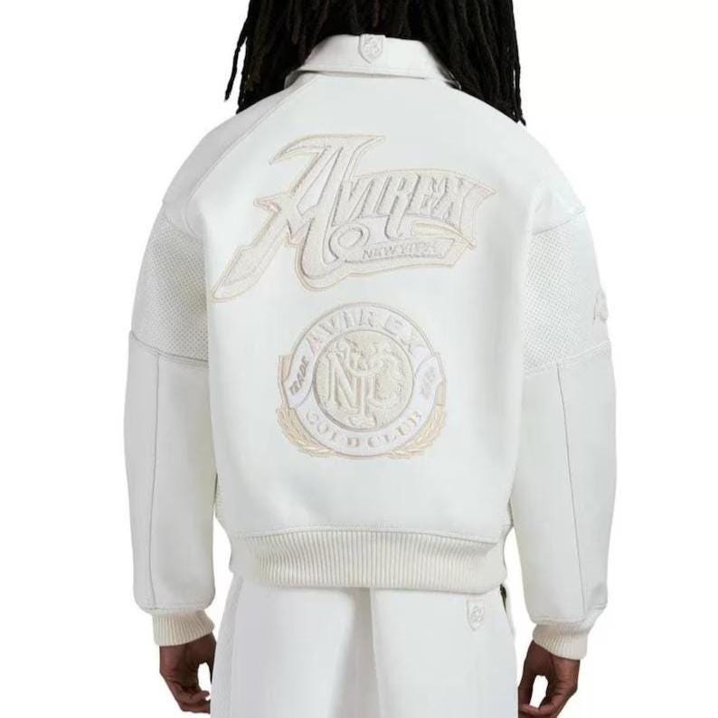 May include: A white varsity jacket featuring a large embroidered Avirex New York logo on the upper back and a circular Avirex Gold Club patch below it. The jacket has a ribbed collar, cuffs, and hem, with textured panels on the sleeves.