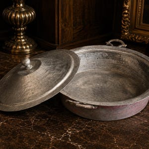 Puede incluir: Una olla antigua de metal martillado con tapa a juego. La olla está abierta, revelando su interior. La tapa descansa junto a la olla. La olla tiene dos asas y un pequeño pomo en la tapa. La olla es de color plateado.